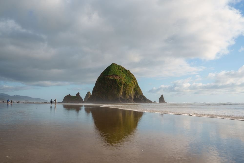 The massive Haystack Rock monolith reflecting on the wet sandy shore at Cannon Beach Oregon under a cloudy sky.