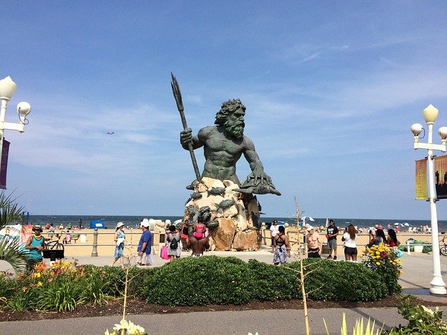 The large bronze King Neptune statue standing near the boardwalk with tourists and the ocean background at Virginia Beach.
